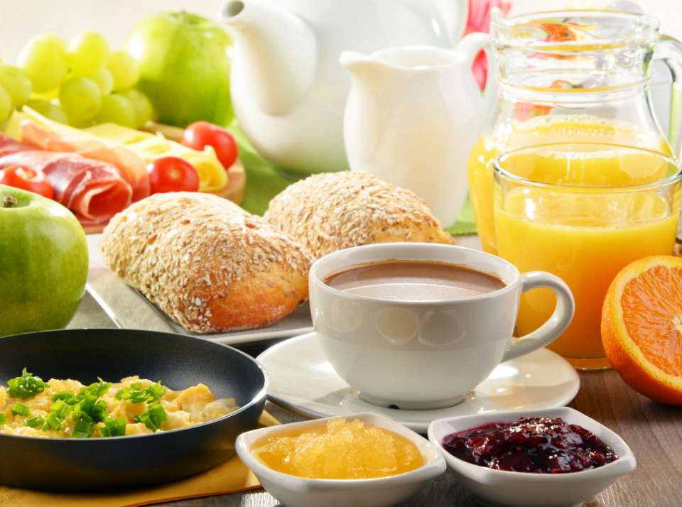 Uma mesa de café da manhã com paes frutas, sucos e uma caneca de GCOFFEE growth supplements