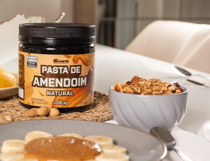 Um ponte de pasta de amendoim 1kg em cima de uma mesa, com uma tigela com grãos na frente dela e um prato com bananas