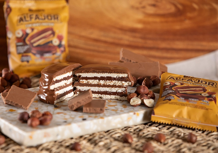 Alfajor com Whey Protein sabor chocolate cortado ao meio em cima de uma tábua com 2 embalagens em volta